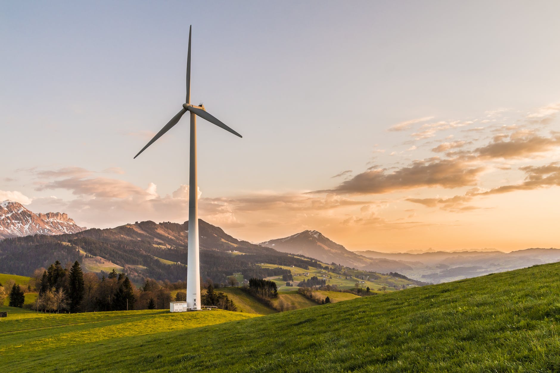 Sunset or dawn-time photo of a white wind turbine positioned in front of a mountainous landscape and green fields. 