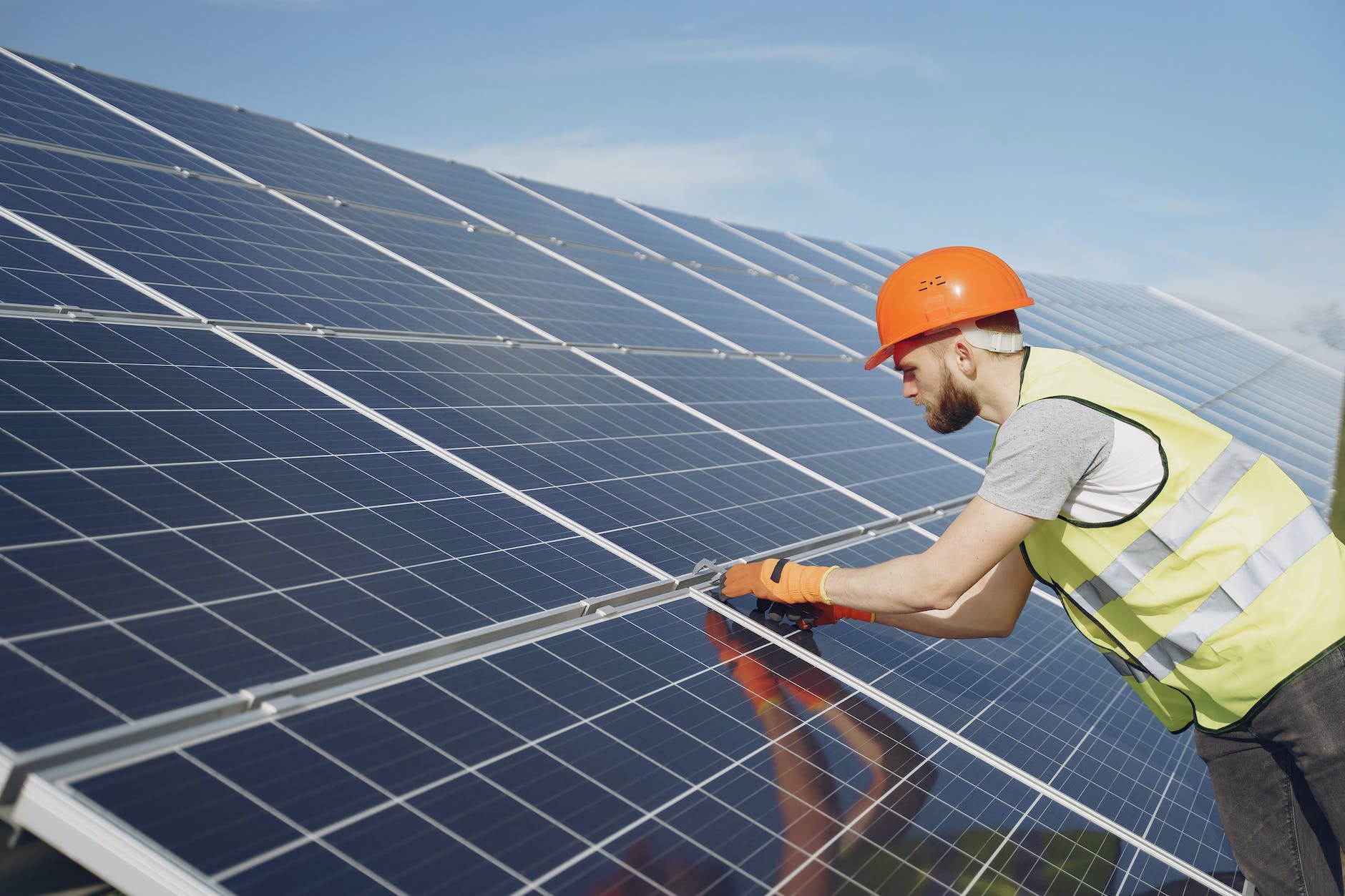 Close shot of a labour worker in a high-visibility jacket and orange helmet working on a large solar panel. 