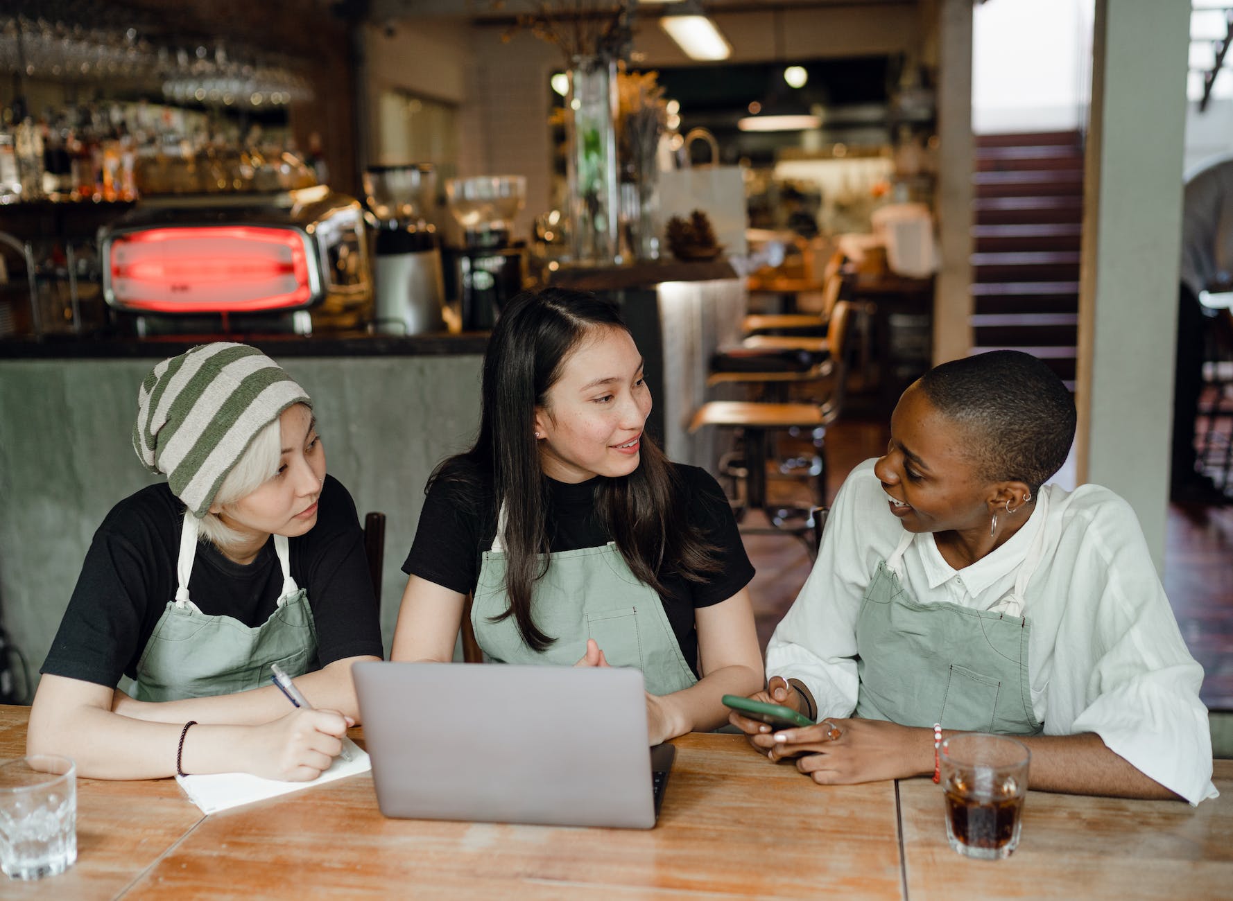 Photo of three people looking at each other, sat at a wooden table wearing green aprons in front of a bar counter top in a restaurant. 